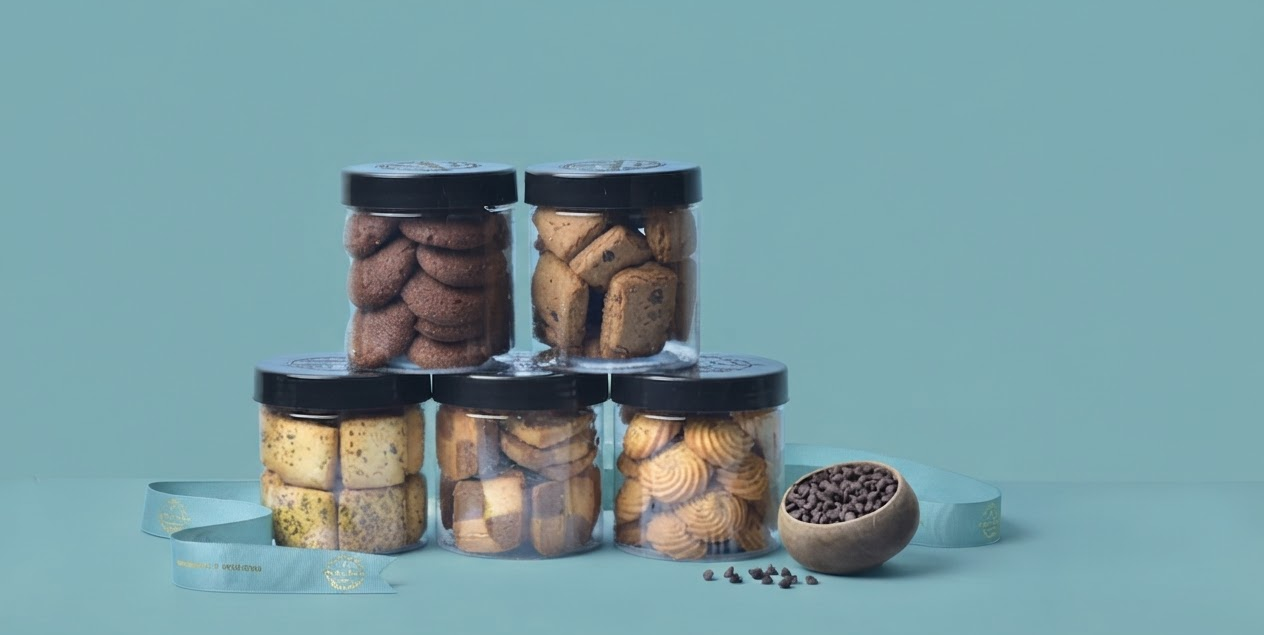 Five jars of Dunkel Braun cookies stacked in a pyramid beside a bowl of chocolate chips and a teal gift ribbon