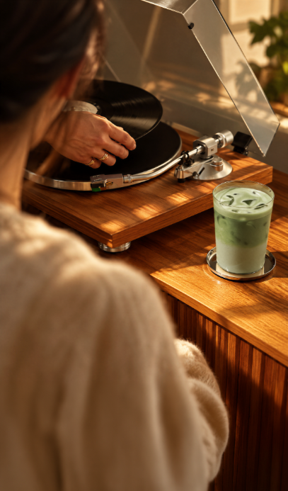 Someone playing a record with a glass of iced pea matcha beside the turntable
