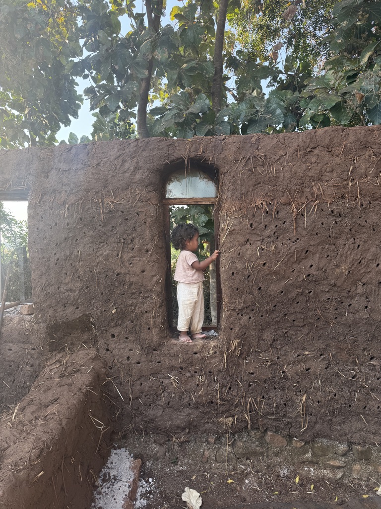 A child standing in the arched window of a hand-built mud wall, framed by trees